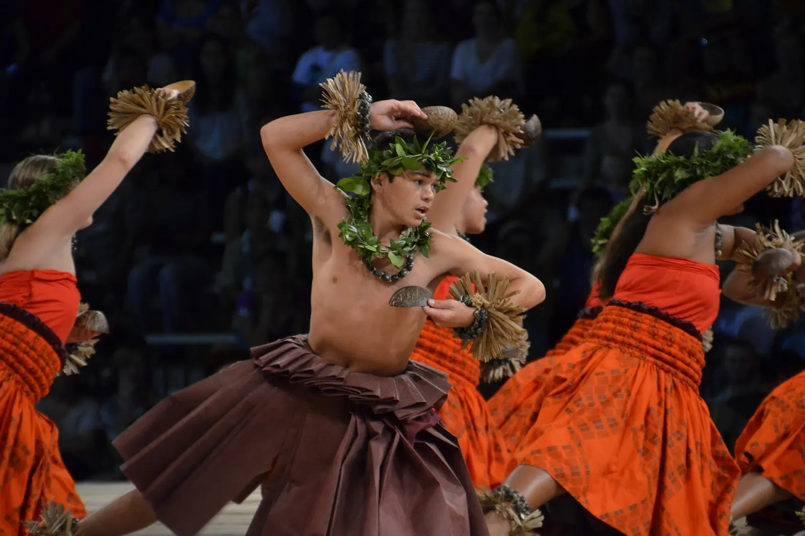 Un bailarín de hula kahiko con una corona de hojas y una falda marrón a mitad de paso sobre el escenario, junto a bailarines con faldas naranjas